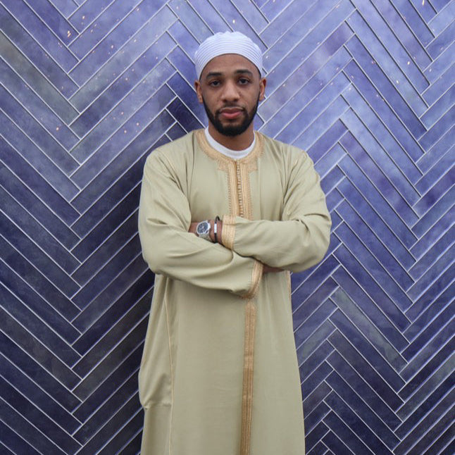 Man with arms folded in a cream Moroccan thobe against a blue tiled wall